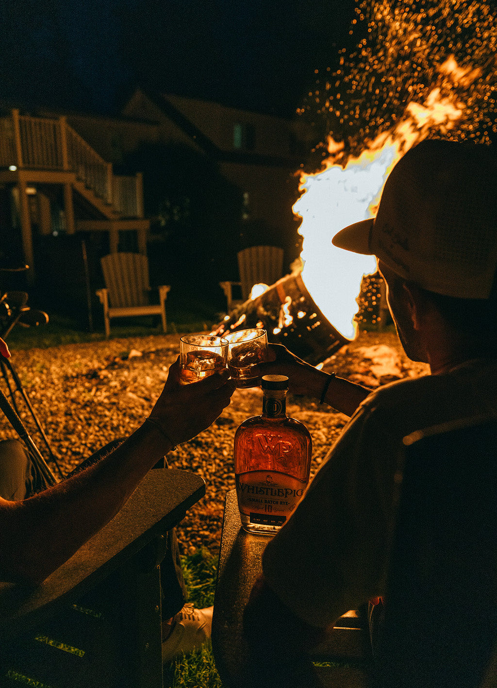 Two people toasting with bottles by a bonfire at night.